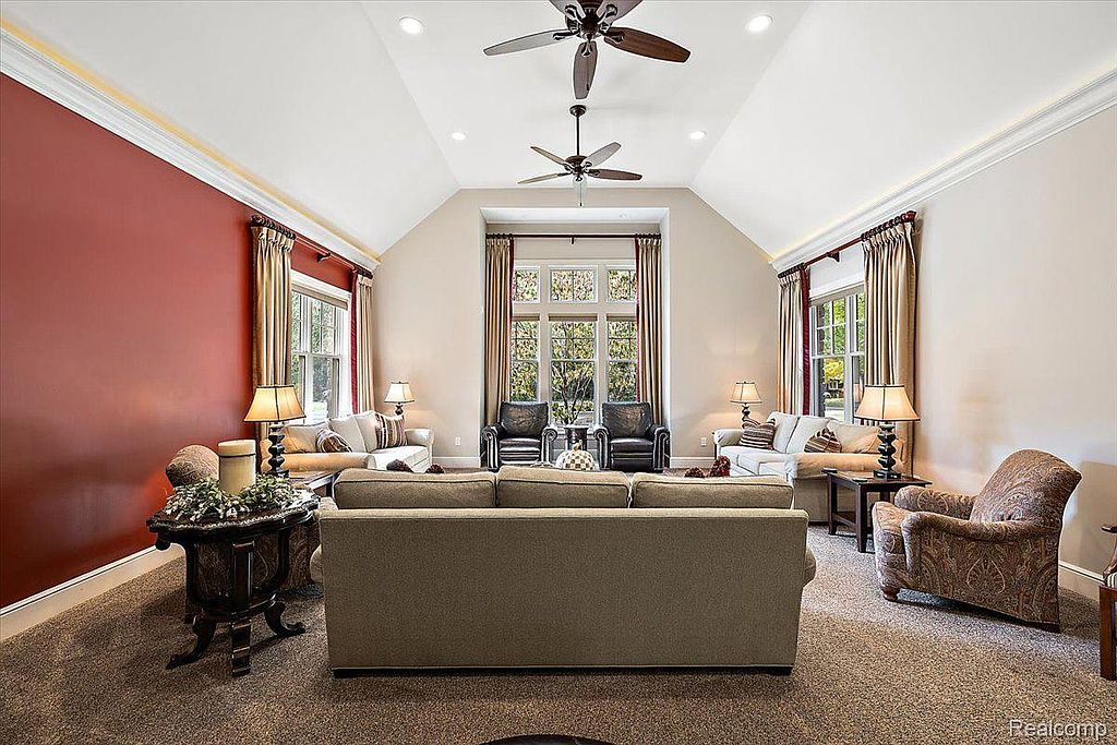 This living room features a high, vaulted ceiling with two ceiling fans and recessed lighting. Large windows with elegant draperies provide natural light and views. The room is furnished with comfortable seating, including sofas and armchairs, arranged around a central area, creating a welcoming and sophisticated atmosphere.