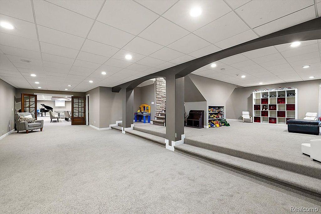 This is a spacious finished basement featuring neutral carpeting and paint, with architectural columns and steps defining different zones. The area includes a play area with toys and shelving, and a separate room visible through double doors, suggesting a versatile space for recreation and relaxation. The overall impression is clean, well-lit, and family-friendly.