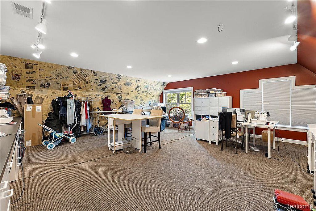 This is a spacious office or study area featuring a unique design with newspaper-covered walls on one side and a red accent wall on the other. The room is well-lit and furnished with multiple desks, chairs, and storage units, suggesting a functional and creative workspace. The overall impression is a blend of eclectic style and practicality.