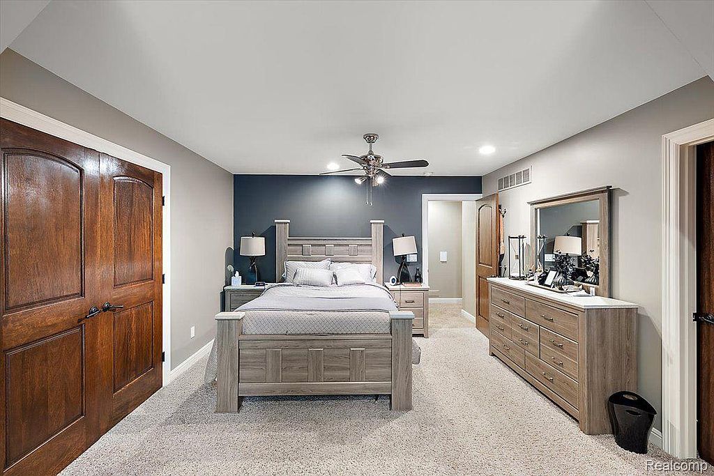 This is a well-lit primary bedroom featuring a large bed with a gray wood frame, matching nightstands, and a dresser with a mirror. The walls are painted in neutral tones, with a darker accent wall behind the bed. The room is carpeted and includes a ceiling fan, creating a comfortable and inviting atmosphere.