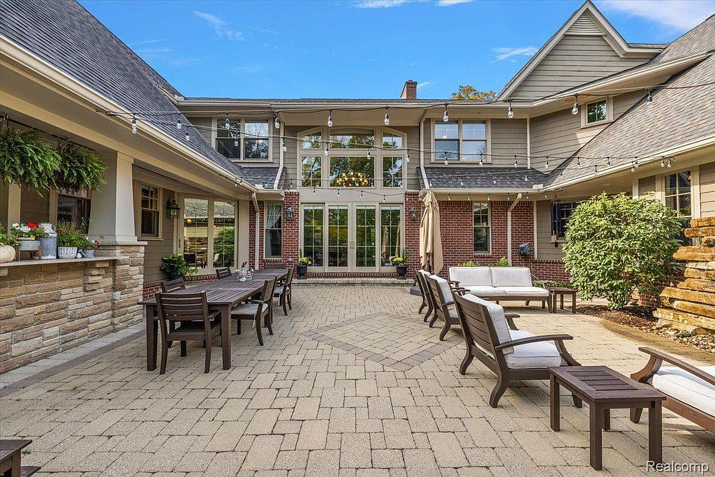 This image showcases a spacious outdoor patio area, perfect for entertaining. The patio features a large dining table with chairs, as well as comfortable lounge seating. The area is surrounded by the home's exterior, which includes brick and siding elements, and is paved with light-colored stones.