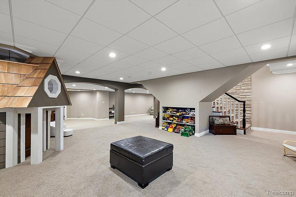 This is a spacious finished basement featuring a play area with a playhouse, a storage unit filled with toys, and a comfortable seating area near the stairs. The room is well-lit with recessed lighting and has neutral-colored carpeting, creating a versatile and inviting space for recreation and relaxation. The overall impression is a clean and family-friendly environment.