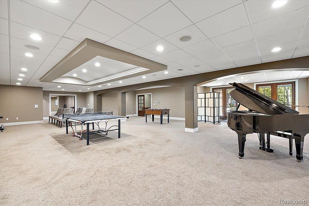 This is a spacious finished basement featuring neutral carpeting and recessed lighting. The area includes a ping pong table, foosball table, and a grand piano, suggesting a recreational space. Arched doorways and columns add architectural interest, while the overall impression is one of a large, versatile entertainment area.
