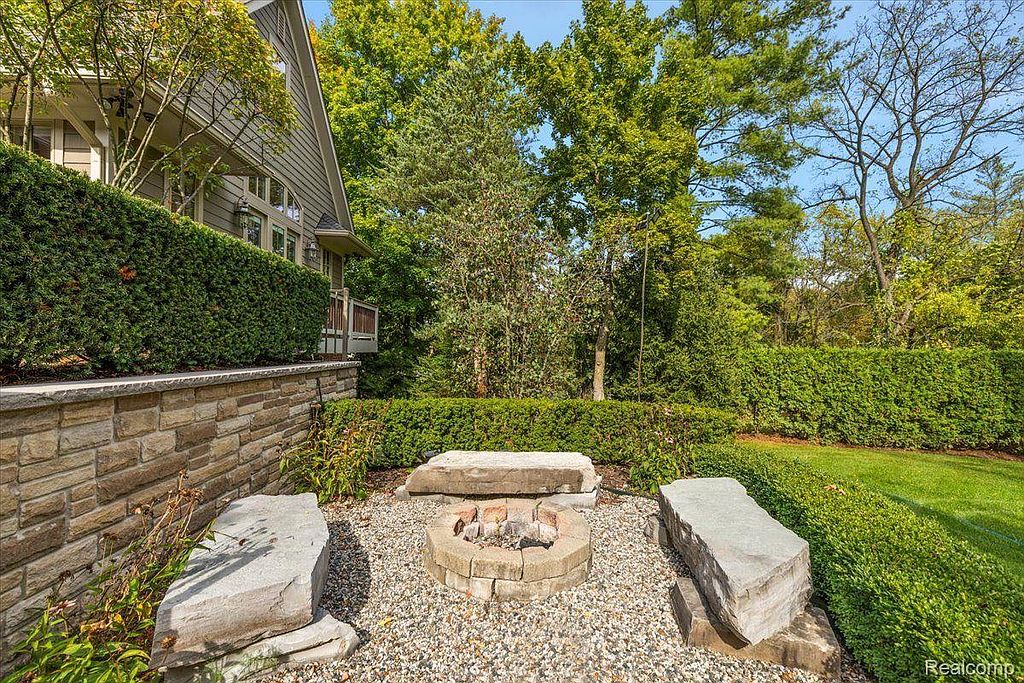 This image showcases a beautifully landscaped backyard featuring a stone fire pit surrounded by gravel and large stone benches. Lush green hedges provide privacy, and mature trees offer shade. The scene creates a cozy and inviting outdoor living space.