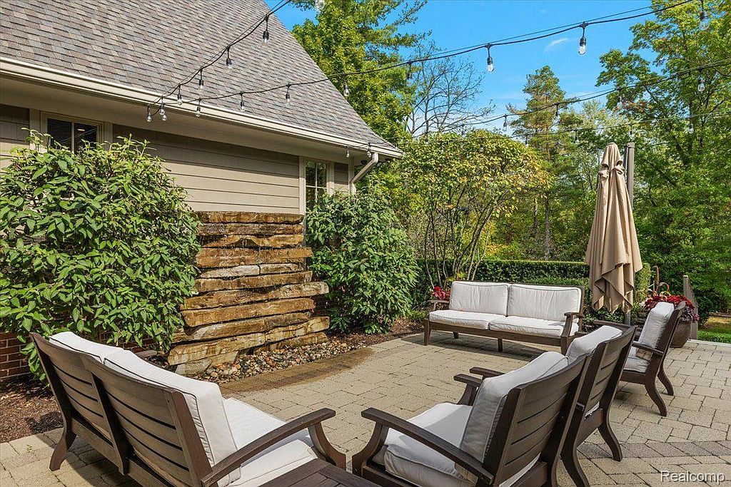 This image showcases a beautifully designed outdoor patio area, perfect for relaxation and entertaining. The space features comfortable seating arrangements, including chairs and a sofa, arranged around a stone water feature. String lights overhead add a warm and inviting ambiance, enhancing the overall appeal of this outdoor living space.