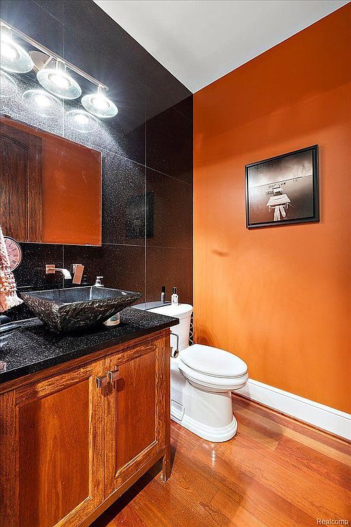 This is a stylish guest bathroom featuring a unique vessel sink on a dark granite countertop and a wooden vanity. The walls are a combination of dark tile and orange paint, complemented by wood flooring. A modern toilet and a framed picture add to the room's sophisticated aesthetic.