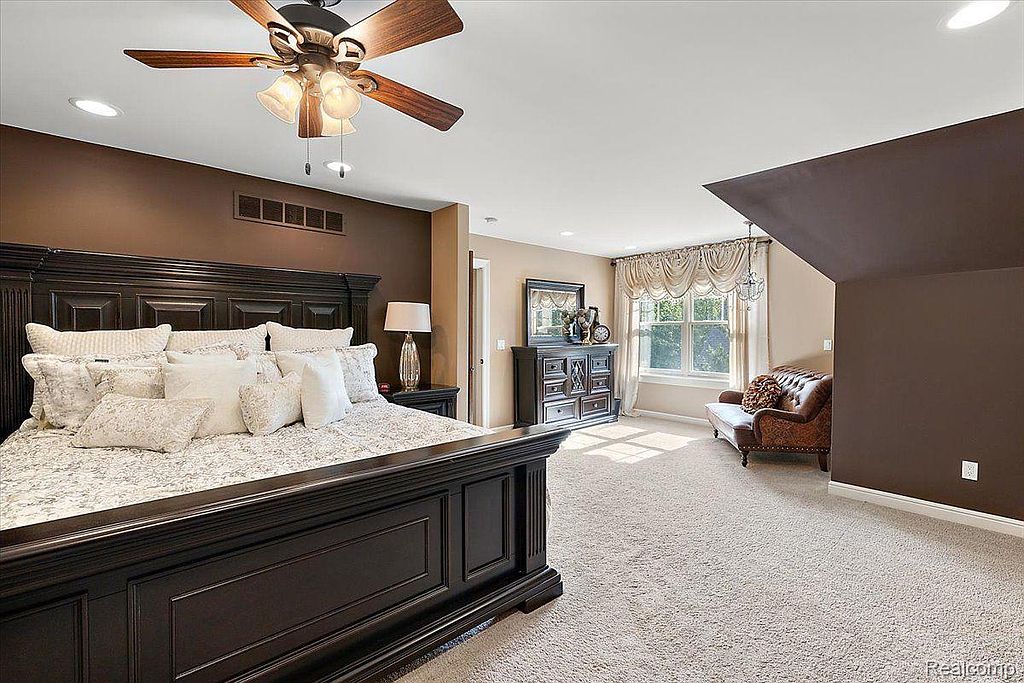 This is a primary bedroom featuring a large, dark wood bed with numerous pillows, creating a luxurious and inviting space. The room has a mix of brown and beige tones, with a sitting area near the window, suggesting a comfortable and relaxing atmosphere. A ceiling fan adds to the room's functionality and style.