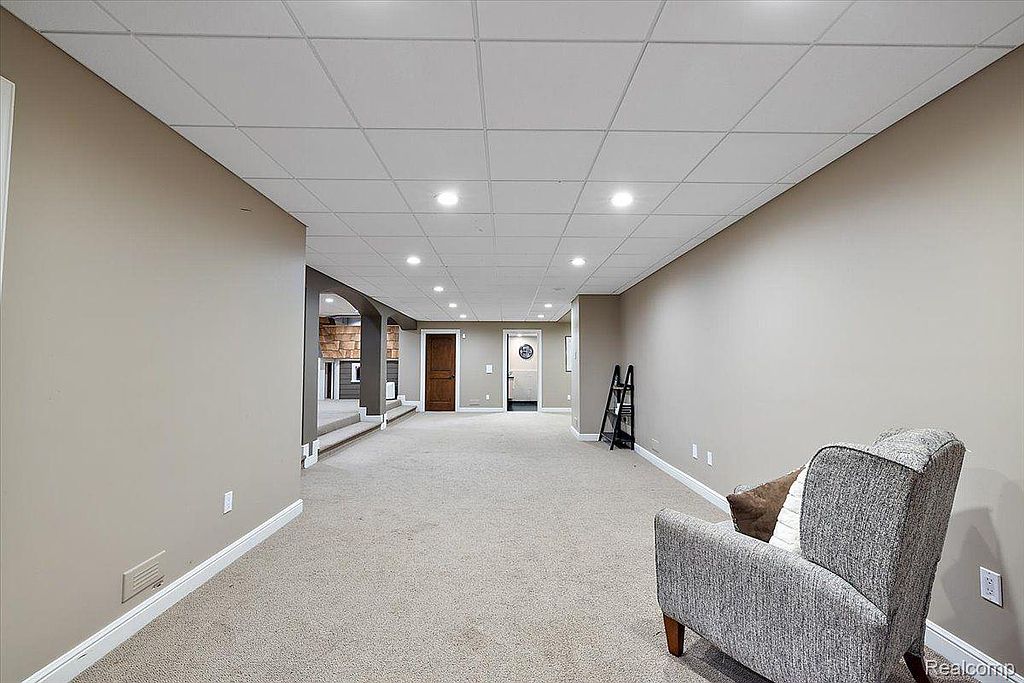 This is a spacious finished basement featuring neutral-toned walls and carpeting. The room has a drop ceiling with recessed lighting, and there's an adjacent area with a fireplace. The overall impression is a clean and versatile space suitable for various uses.
