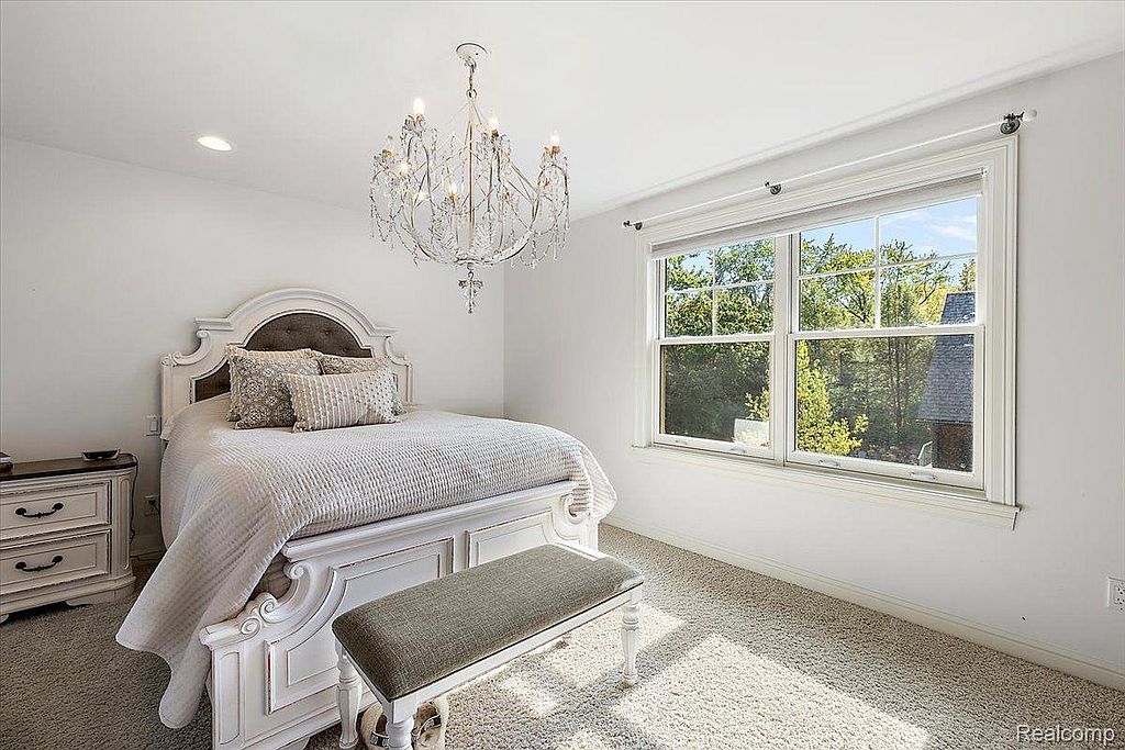 This is a bright and airy primary bedroom featuring a large bed with a decorative headboard and neutral-toned bedding. A crystal chandelier adds a touch of elegance, while a large window provides natural light and a view of the outdoors. The room is decorated in a clean, classic style with white walls and light-colored carpeting.