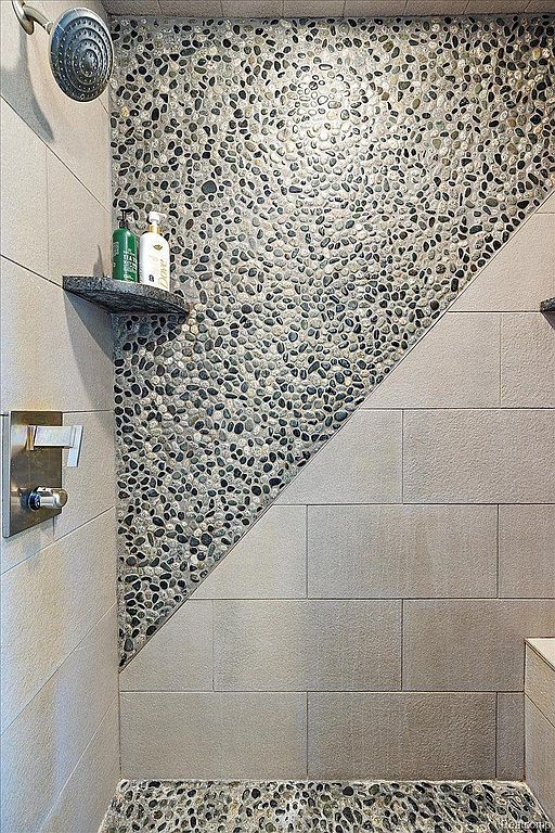 This image showcases a stylish bathroom with a unique pebble-stone accent wall and floor, complemented by large rectangular tiles. A built-in shelf holds toiletries, and the shower fixtures are modern and sleek. The design creates a spa-like atmosphere, emphasizing natural textures and a clean aesthetic.