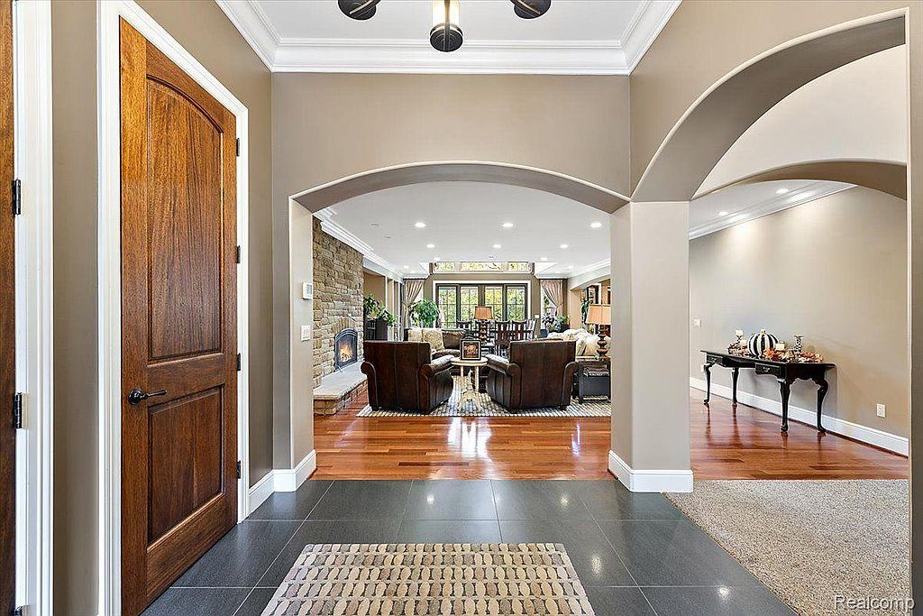 This image showcases a grand entryway featuring dark tile flooring transitioning to hardwood, arched doorways, and a view into a spacious living area with a stone fireplace. The warm color palette and architectural details create an inviting and luxurious atmosphere, highlighting the home's elegant design and flow.