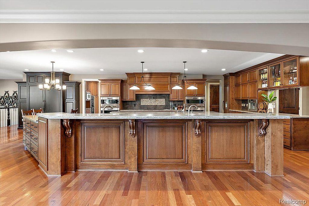 This is a spacious kitchen featuring wooden cabinetry, granite countertops, and a large central island with decorative corbels. The kitchen is well-lit with recessed lighting and pendant lights over the island, creating a warm and inviting atmosphere. The hardwood flooring adds to the overall elegance of the space.