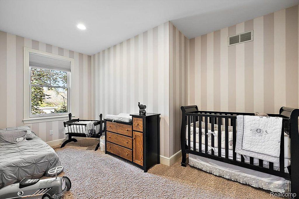 This is a charming nursery featuring striped wallpaper in neutral tones. The room includes a crib, a changing table dresser, and a child's bed with a toy car, creating a cozy and inviting space for a young child. The overall impression is neat and well-decorated.