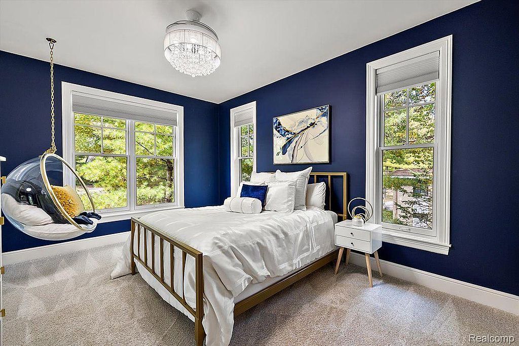 This is a stylish bedroom featuring a navy blue accent wall, a brass-framed bed with white linens, and a unique hanging bubble chair. Natural light streams in through multiple windows, and a modern chandelier adds a touch of elegance. The room has a contemporary and inviting feel.