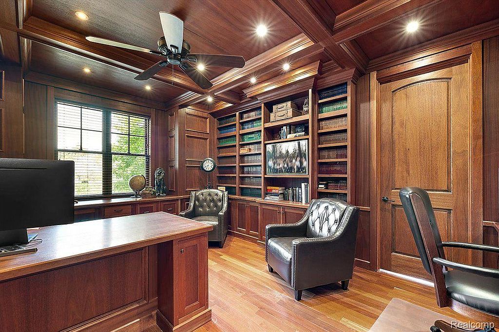This is a luxurious home office featuring rich wood paneling, built-in bookshelves, and hardwood floors. The room is furnished with leather armchairs and a large desk, creating a sophisticated and comfortable workspace. A ceiling fan and recessed lighting provide ample illumination.