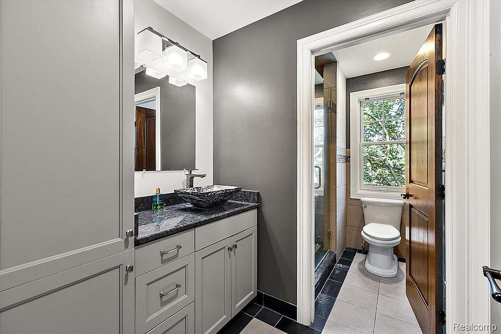 This bathroom features a modern vanity with a unique vessel sink and granite countertop. The gray walls and cabinetry create a sleek, contemporary look. A doorway leads to a separate toilet and shower area, enhancing the functionality of the space.