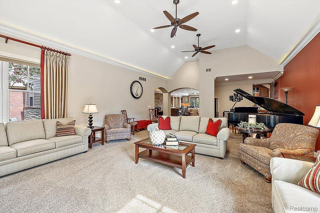 This is a spacious living room featuring neutral-toned sofas, a dark wood coffee table, and a grand piano in the background. The room has high ceilings with ceiling fans and recessed lighting, creating a bright and airy atmosphere. The overall impression is one of elegance and comfort, suitable for both relaxation and entertaining.