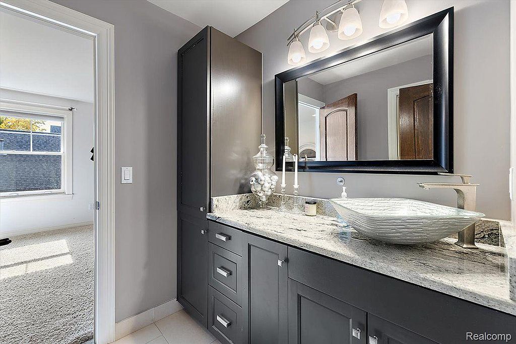 This is a primary bathroom featuring a modern vanity with a unique vessel sink and a large, framed mirror. The color palette is neutral, with gray cabinetry and walls, complemented by a light-colored countertop. The bathroom appears clean and well-maintained, offering a luxurious and functional space.