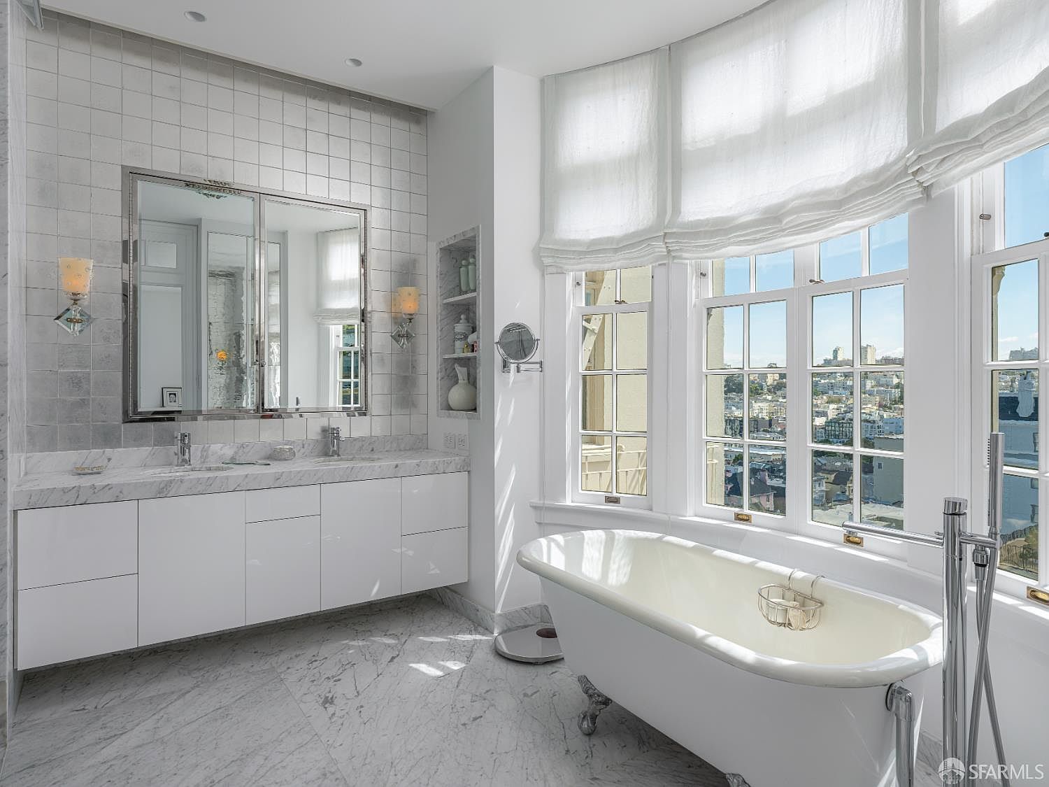 This bright and elegant primary bathroom features a classic clawfoot tub positioned beneath large windows that offer an expansive city view. The space is finished with light-toned stone flooring and wall tiles, complemented by a sleek, modern double vanity with a stone countertop and a large, framed mirror. The room is bathed in natural light, creating a clean, sophisticated, and airy atmosphere.