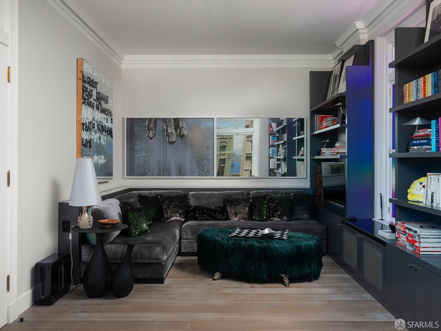 This contemporary living room features a plush, dark grey sectional sofa adorned with shimmering accent pillows, paired with a vibrant, deep green faux-fur ottoman. The space is framed by dark, built-in shelving units and a large, multi-panel wall art installation that reflects the room's sophisticated, moody aesthetic. Light wood-toned flooring provides a neutral base, while a modern lamp and side tables complete this stylish, intimate lounge area.