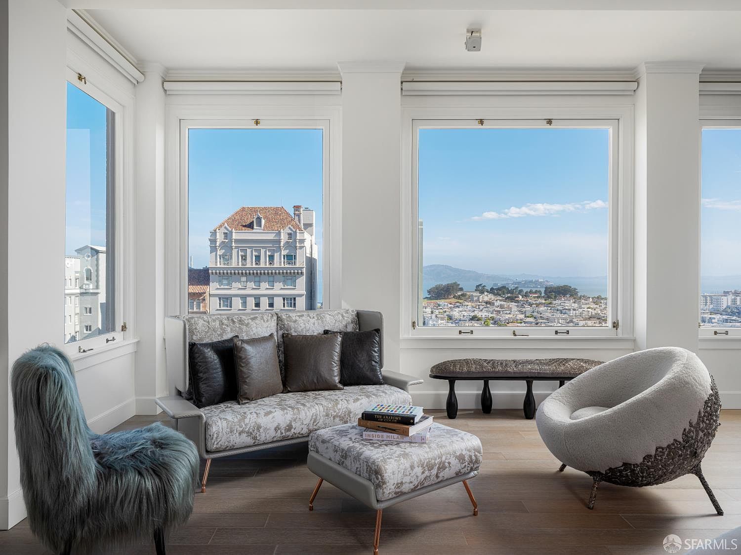 This sophisticated living area features a curated selection of modern furniture, including a patterned sofa, a matching ottoman, a textured blue armchair, and a sculptural, rounded lounge chair. Large windows frame expansive, elevated views of the city and the bay, bathing the room in natural light. The space is finished with light wood flooring and a minimalist aesthetic, creating an airy and refined atmosphere.
