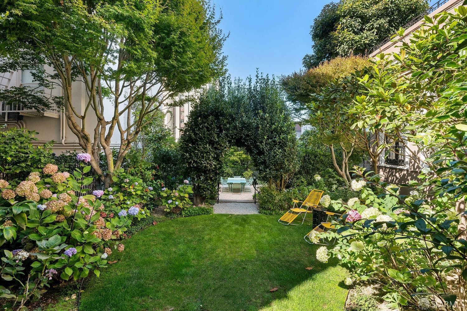 This lush, private garden features a well-manicured lawn framed by vibrant hydrangea bushes and mature trees. A central arched hedge creates a charming focal point, leading to a secluded patio area with an outdoor dining set. The perspective is a straight-on view from the lawn, capturing the serene and verdant atmosphere of this tranquil outdoor space.