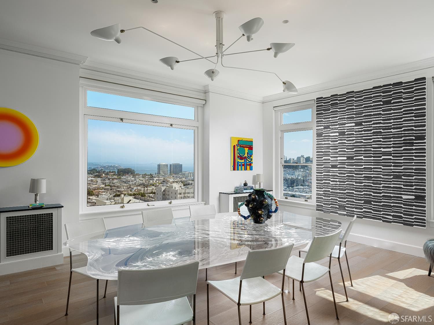 This bright dining room features a unique, translucent, fluid-shaped table surrounded by modern white chairs. The space is anchored by a sculptural, multi-armed chandelier and large windows that frame an expansive city and bay view. Artful decor, including a vibrant abstract painting and a large, textured black-and-white wall piece, adds a sophisticated, gallery-like aesthetic to the room.