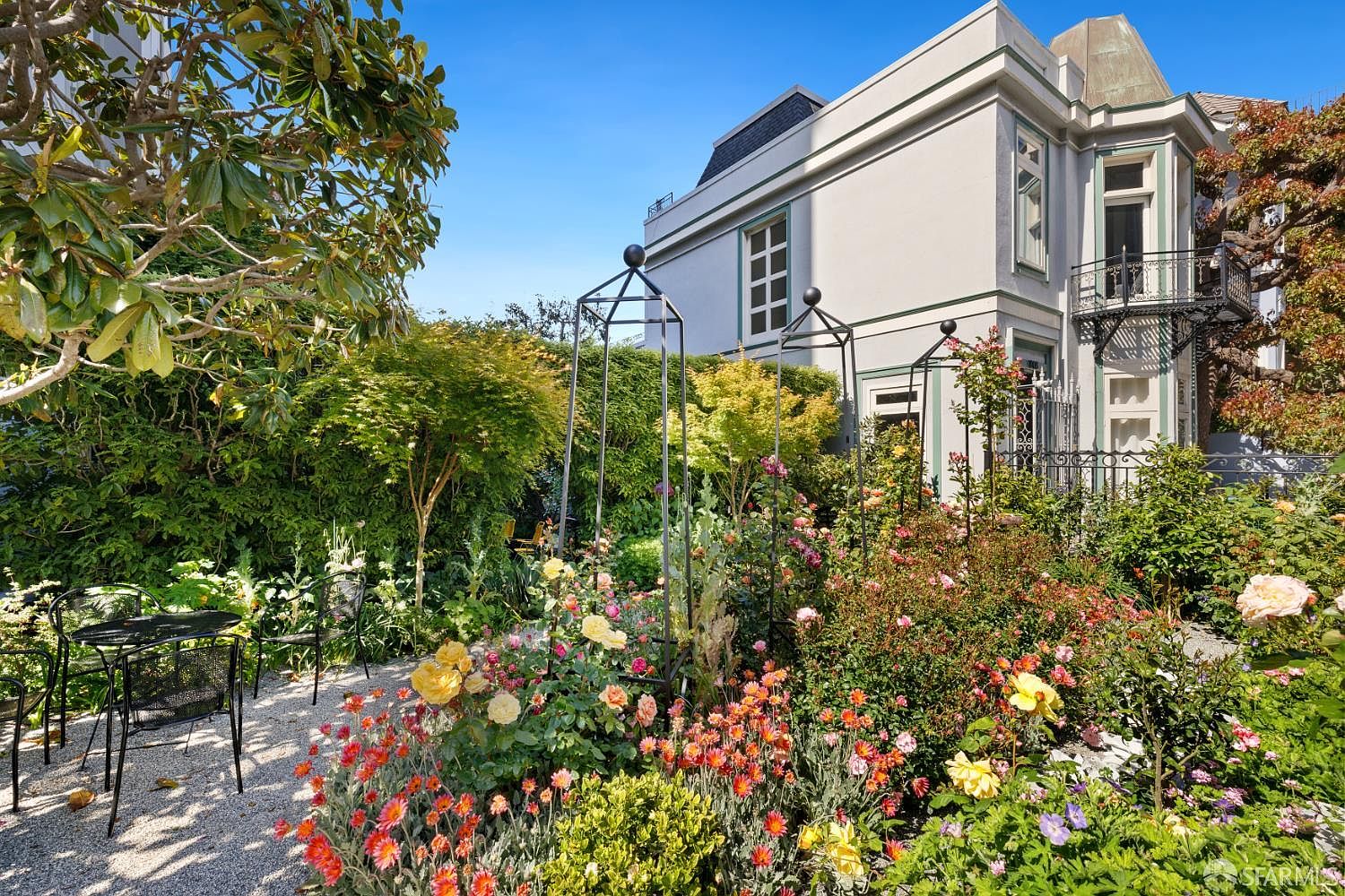 This lush, vibrant garden features a variety of blooming flowers, including yellow roses and orange daisies, set against a backdrop of mature trees and a manicured hedge. A small bistro set sits on a gravel patio area, providing a peaceful spot to enjoy the scenery. The scene is framed by the side of a classic, light-colored home with elegant architectural details and a decorative wrought-iron balcony.