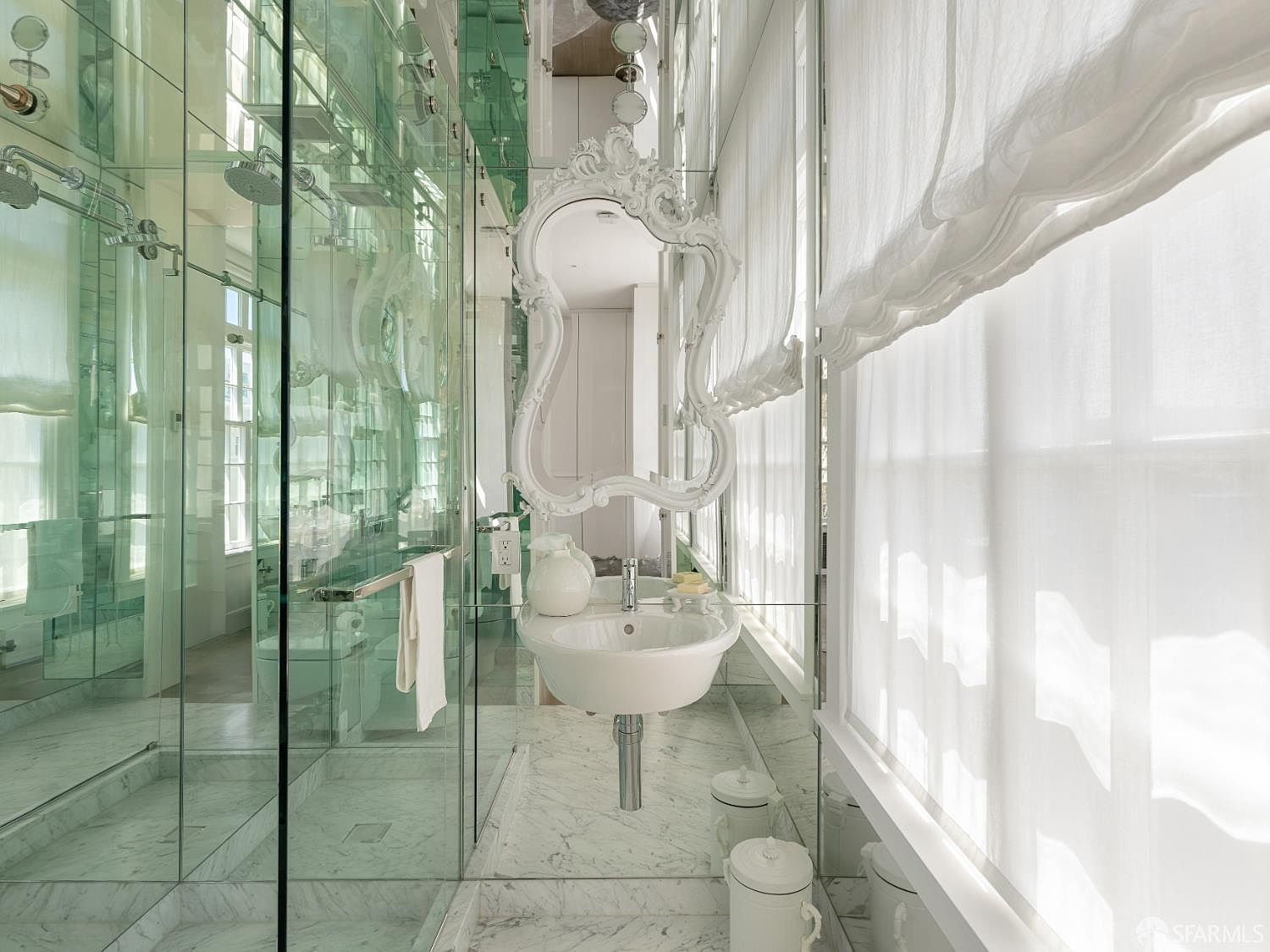 This luxurious bathroom features extensive mirrored walls that create a sense of infinite space, complemented by a white, ornate-framed mirror above a wall-mounted pedestal sink. A glass-enclosed shower area with stone flooring and walls adds to the sleek, reflective aesthetic, while sheer white curtains soften the light from the large windows. The overall impression is one of high-end, sophisticated design with a bright and airy atmosphere.