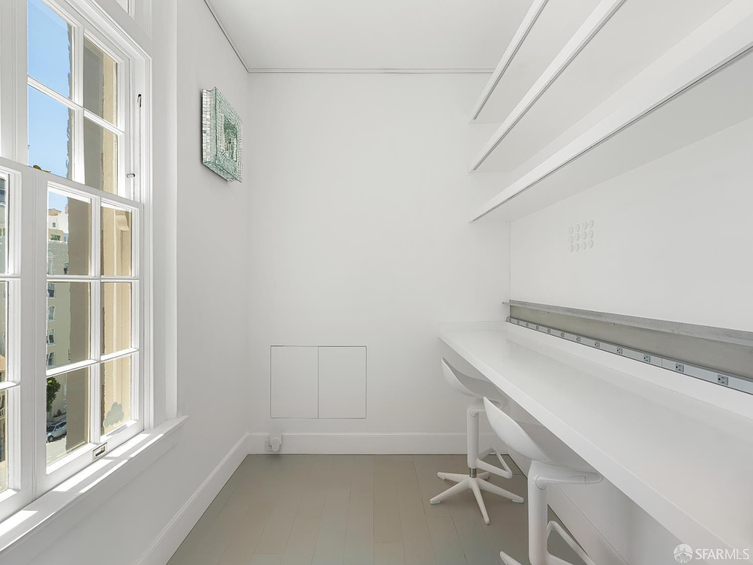 This minimalist home office or study area features a long, built-in white desk surface paired with two modern white chairs. The room is bright and airy, highlighted by a large window that allows for ample natural light, and includes open shelving mounted on the wall above the desk. The space is finished with light-toned flooring and clean white walls, creating a functional and uncluttered workspace.