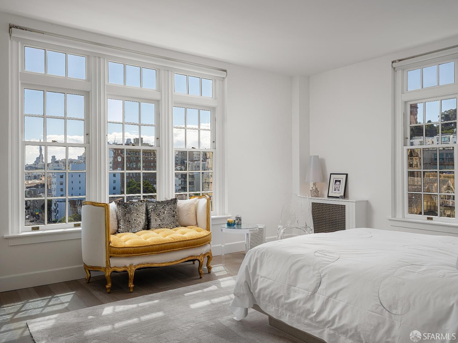 This bright and airy primary bedroom features a classic aesthetic with white walls and light wood flooring. A distinctive gold-framed settee with yellow upholstery sits beneath a large bank of windows that offer an expansive city view. The room is furnished with a white-covered bed, a clear acrylic chair, and a small glass side table, creating a sophisticated and serene atmosphere.