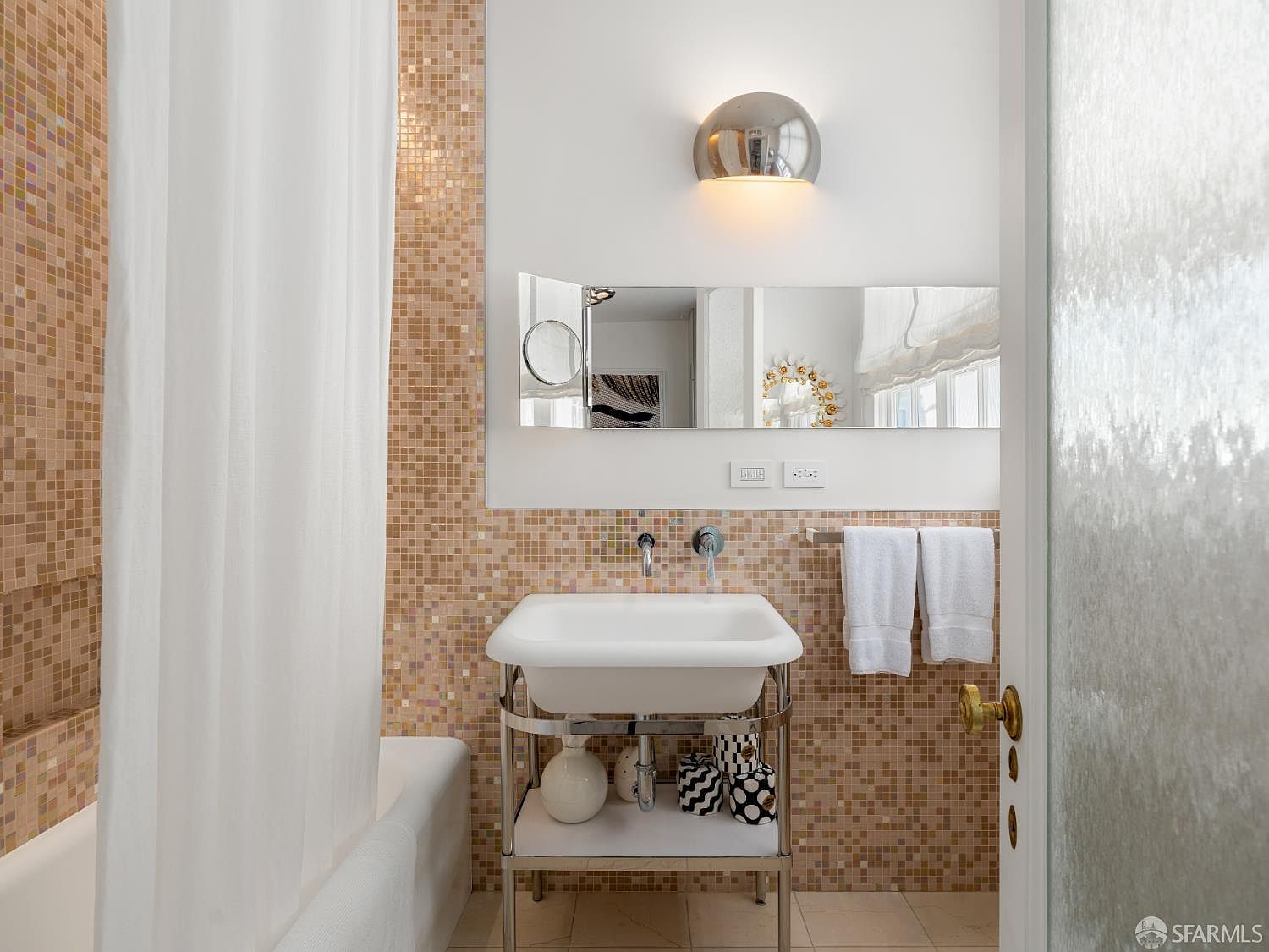 This compact bathroom features a pedestal-style sink with a metal frame and a wall-mounted faucet, set against a wall covered in small, warm-toned mosaic tiles. A wide, rectangular mirror is positioned above the sink, complemented by a modern, semi-circular wall sconce. To the left, a white shower curtain hangs beside a bathtub, while two white towels are neatly folded on a rack to the right.