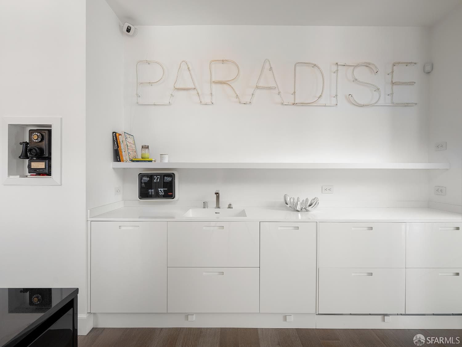 This modern wet bar or kitchenette area features sleek, white handleless cabinetry and a clean white countertop with an integrated sink. Above the counter, a floating shelf holds a digital clock and decorative items, while the wall is adorned with a large, illuminated 'PARADISE' sign. To the left, a recessed wall niche displays a vintage camera, adding a unique, artistic touch to the minimalist space.