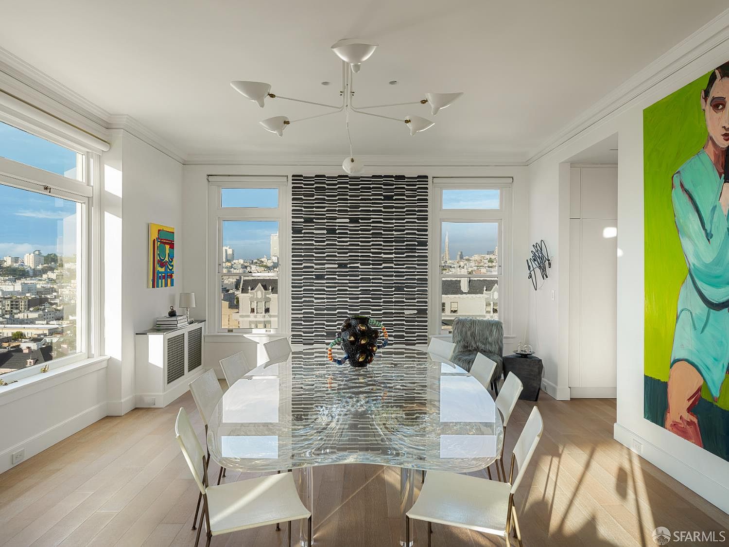 This sophisticated dining room features a unique, sculptural glass table as its centerpiece, surrounded by modern white chairs. The space is anchored by a striking, textured black-and-white wall installation between two large windows that frame expansive city views. A contemporary white chandelier hangs above, while a vibrant, large-scale portrait painting adds a bold pop of color to the light-filled, minimalist interior.