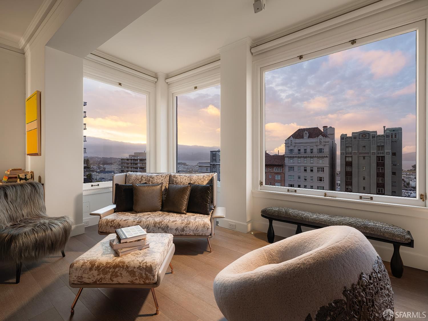 This sophisticated living room features a contemporary seating arrangement with a patterned sofa, a matching ottoman, and a plush, rounded armchair. Large windows frame a stunning panoramic view of the city skyline and bay at sunset, bathing the room in warm, natural light. The space is finished with light wood flooring and a minimalist aesthetic, creating an elegant and serene atmosphere.