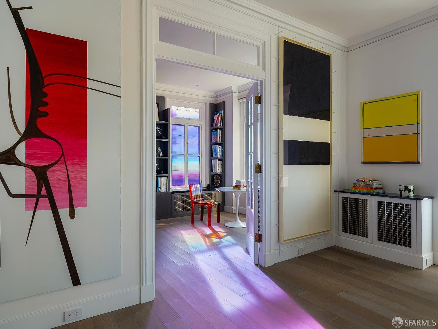 This view captures a transition space between two rooms, characterized by a vibrant, artistic aesthetic and high-contrast decor. A large, abstract red and black painting hangs on the left wall, while a tall, minimalist black and white piece is mounted on the right, flanking a doorway that leads into a sunlit office. The office features a colorful chair and a desk, with light streaming through a window to cast a dramatic, prismatic purple glow across the light wood flooring.