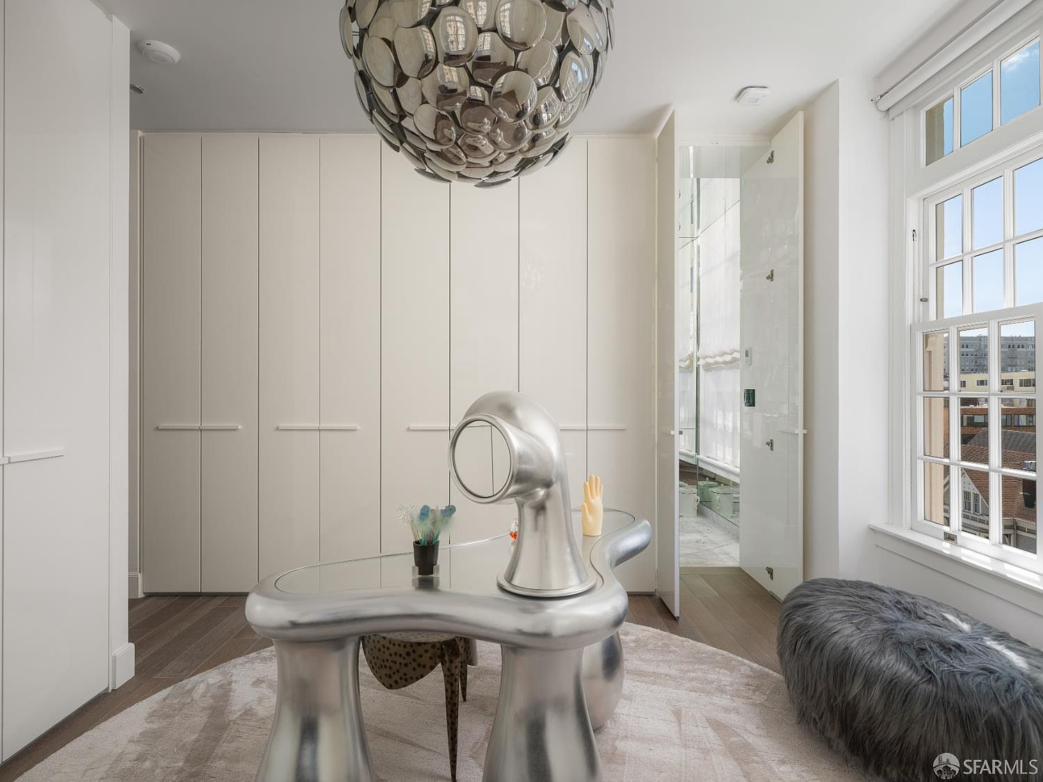 This modern, avant-garde office space features a unique, sculptural silver desk as its centerpiece, positioned on a circular area rug. A wall of floor-to-ceiling cabinetry provides ample storage, while a striking, multi-faceted orb chandelier hangs above. To the right, a large window allows natural light to fill the room, complemented by a plush, grey textured ottoman.