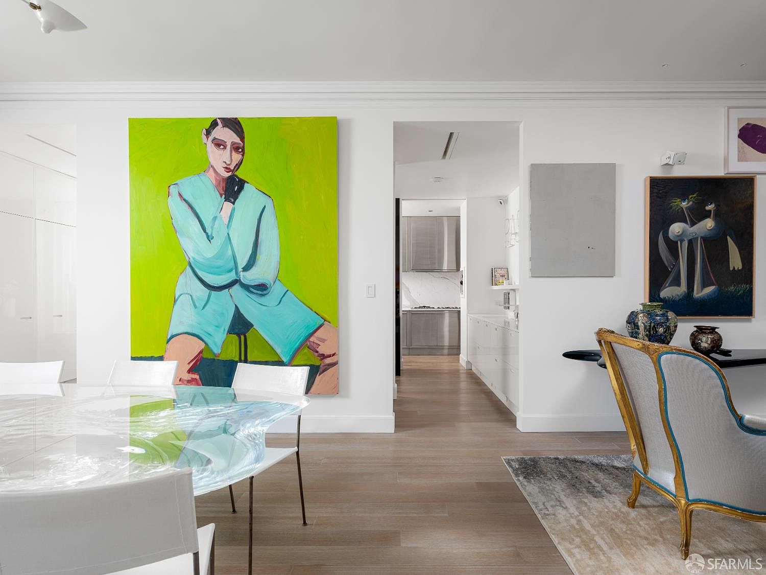 This sophisticated dining area features a unique, sculptural glass table paired with modern white chairs, set against a backdrop of vibrant contemporary art. The space flows seamlessly into a bright, minimalist kitchen visible through a wide doorway, highlighting an open-concept layout with light wood flooring. The room is accented by a classic upholstered chair and a sleek console table, creating a refined and artistic atmosphere.