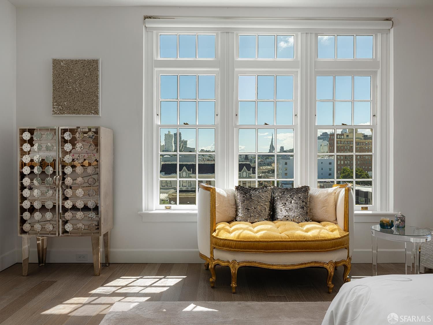 This elegant bedroom space features a luxurious, gold-framed settee with yellow tufted upholstery positioned in front of a large, multi-pane window that offers an expansive city view. To the left, a unique cabinet with a mirrored, circular-patterned facade adds a touch of modern glamour, while light wood flooring grounds the room. The bright, airy atmosphere is enhanced by the natural light streaming through the windows, creating a sophisticated and serene retreat.