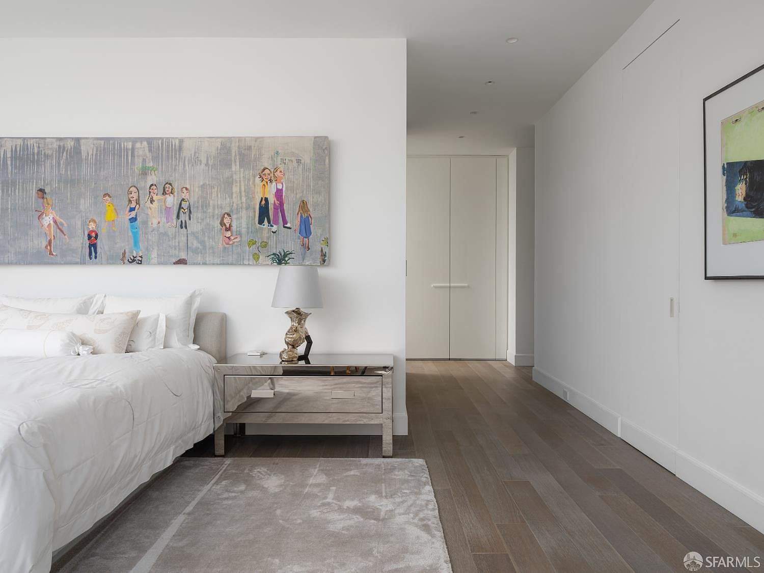 This primary bedroom features a minimalist aesthetic with crisp white walls, light wood flooring, and a large, colorful figurative painting above the bed. A mirrored nightstand with a contemporary lamp sits beside the bed, which is dressed in white linens, while a soft area rug adds texture to the space. The room opens into a hallway with clean, white doors, creating a bright and airy atmosphere.