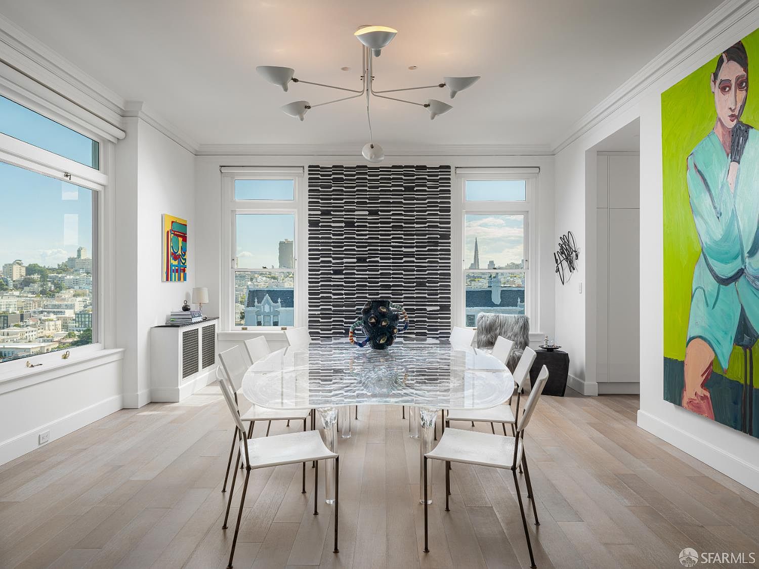 This sophisticated dining room features a large, transparent acrylic table surrounded by sleek, modern chairs, centered beneath a contemporary multi-arm chandelier. The space is defined by light wood flooring, a striking black-and-white textured wall feature, and large windows that frame expansive city views. A vibrant, large-scale portrait painting adds a bold pop of color to the minimalist, gallery-like aesthetic.