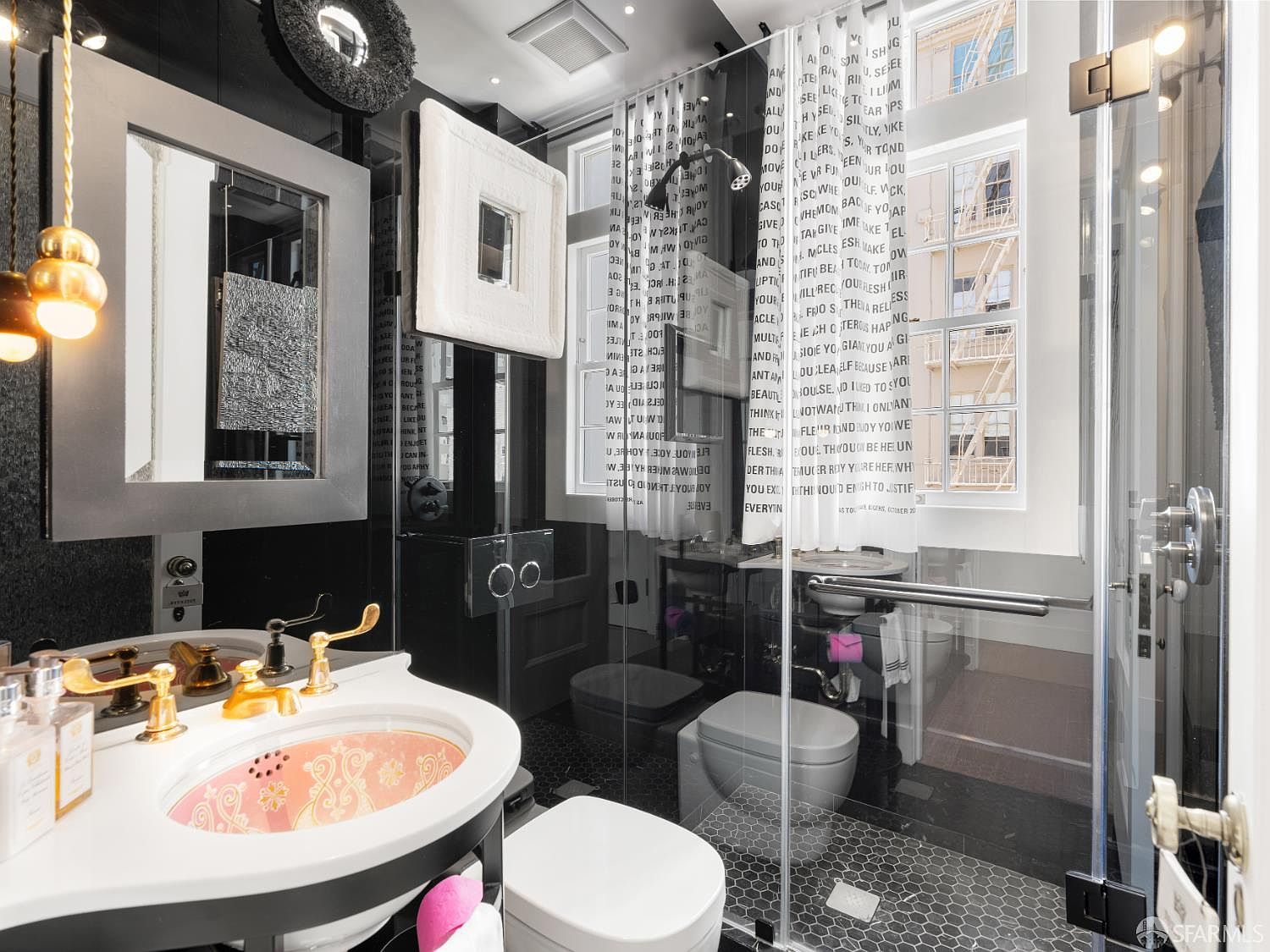 This eclectic bathroom features a unique vanity with a patterned sink basin and gold-toned fixtures, set against a dark, moody backdrop. A glass-enclosed shower stall is adorned with a custom curtain featuring printed text, while the floor is finished in small, dark hexagonal tiles. The space is illuminated by a cluster of spherical pendant lights, creating a bold and artistic aesthetic.