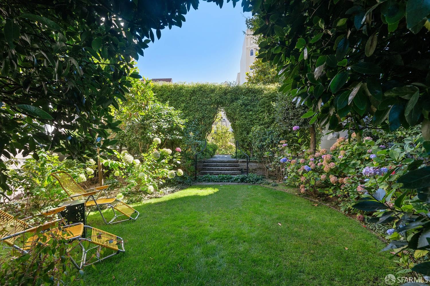 This lush, private backyard features a well-manicured lawn framed by vibrant hydrangea bushes and mature greenery. A set of stone steps leads through a natural archway in the hedge, creating a charming focal point at the rear of the garden. Two yellow lounge chairs are positioned on the grass, offering a peaceful spot to enjoy the serene, sunlit outdoor space.