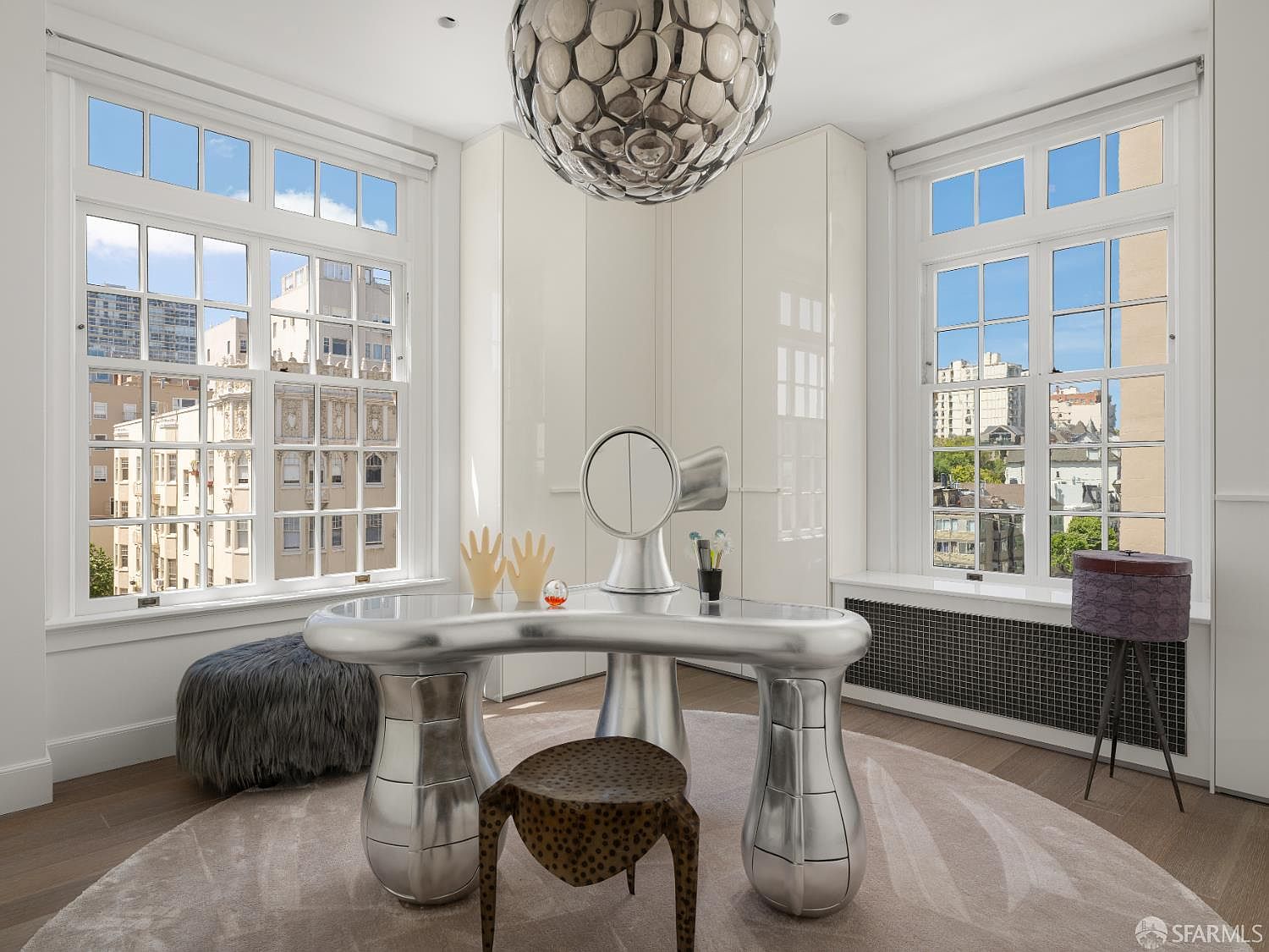This unique office space features a futuristic, metallic desk as the centerpiece, complemented by a sculptural pendant light and a patterned stool. The room is framed by two large windows that provide abundant natural light and views of the surrounding cityscape. Built-in white cabinetry provides clean, minimalist storage, while a textured rug and a plush grey ottoman add softness to the modern aesthetic.