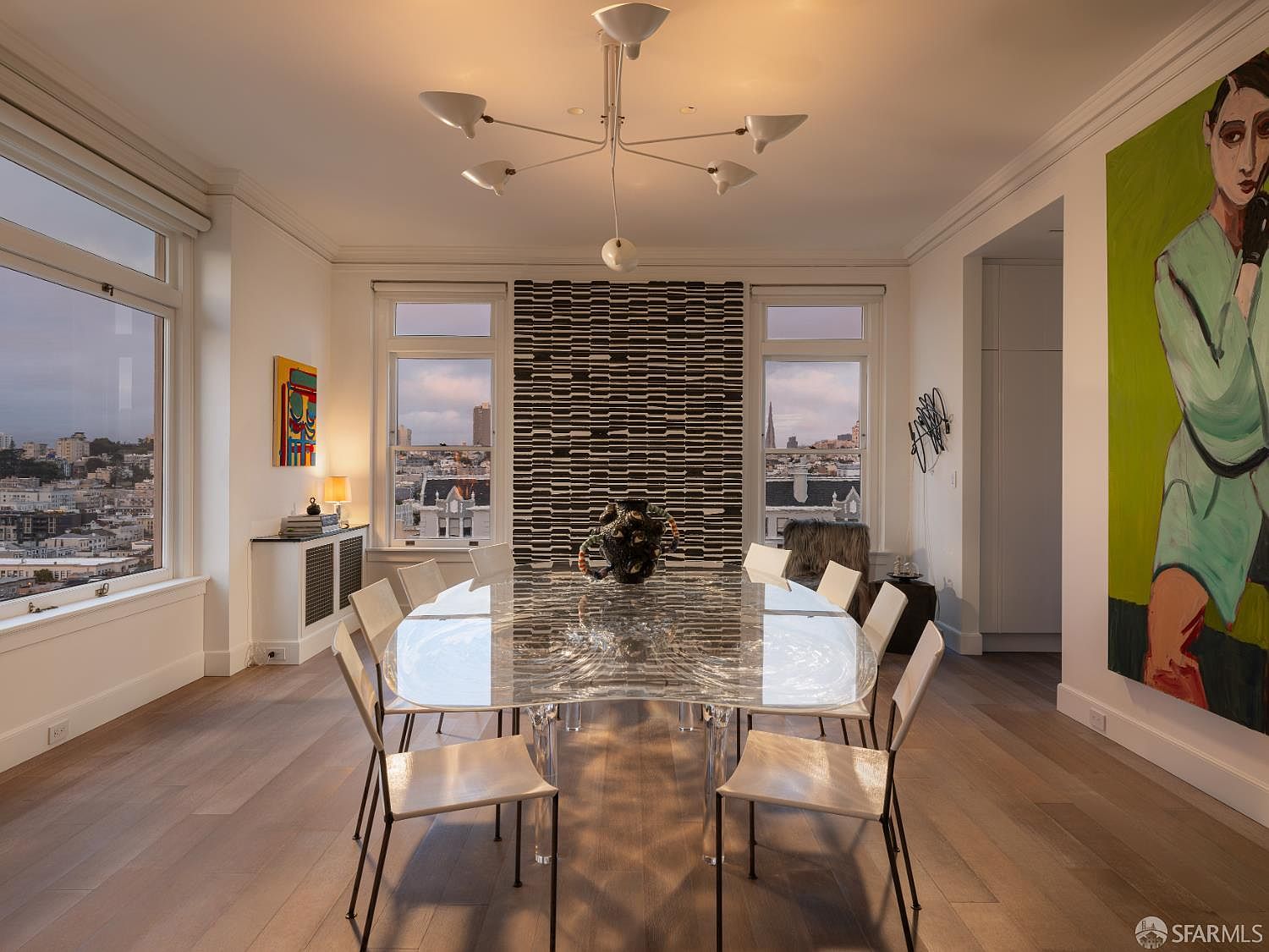 This sophisticated dining room features a unique, sculptural glass-topped table surrounded by eight modern chairs, centered beneath a contemporary multi-arm chandelier. The room is anchored by a striking, textured dark accent wall positioned between two large windows that frame an expansive city view. A large, vibrant piece of figurative art adds a bold splash of color to the otherwise neutral, light-filled space with light wood flooring.