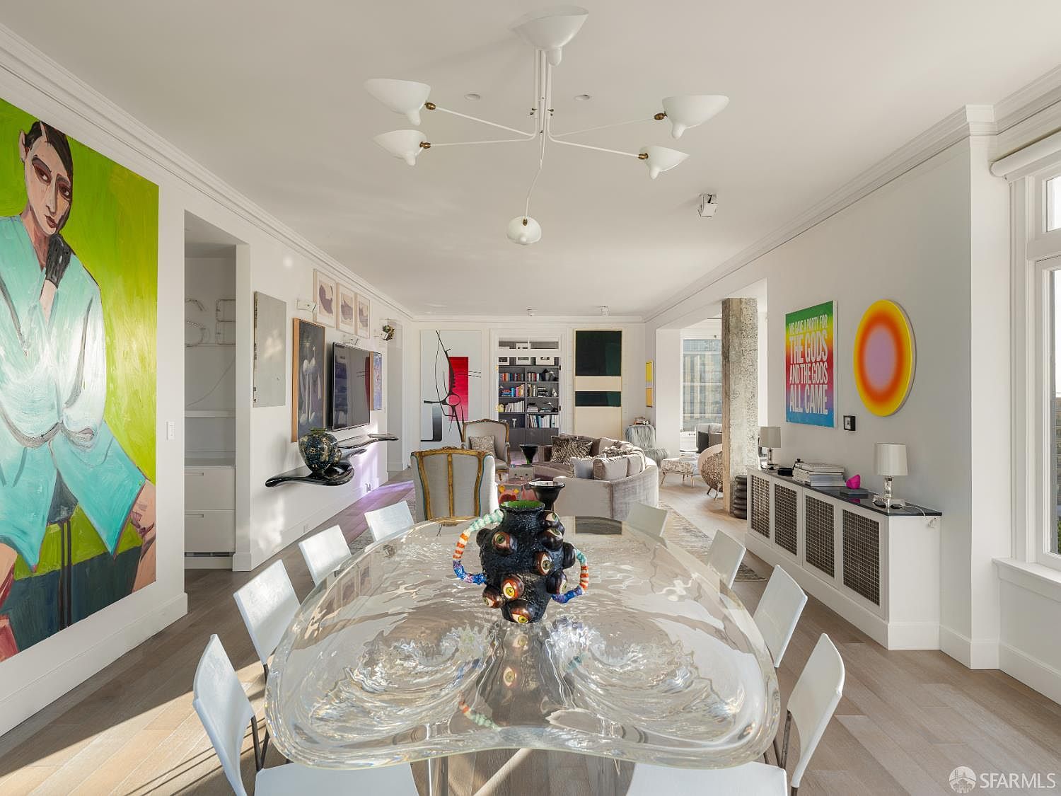 This sophisticated dining area features a unique, sculptural glass table as its centerpiece, surrounded by modern white chairs. The room is decorated with vibrant, large-scale contemporary artwork and a multi-armed chandelier, creating a gallery-like atmosphere. The open-concept layout flows seamlessly into a living space, showcasing light wood flooring and a bright, airy aesthetic.