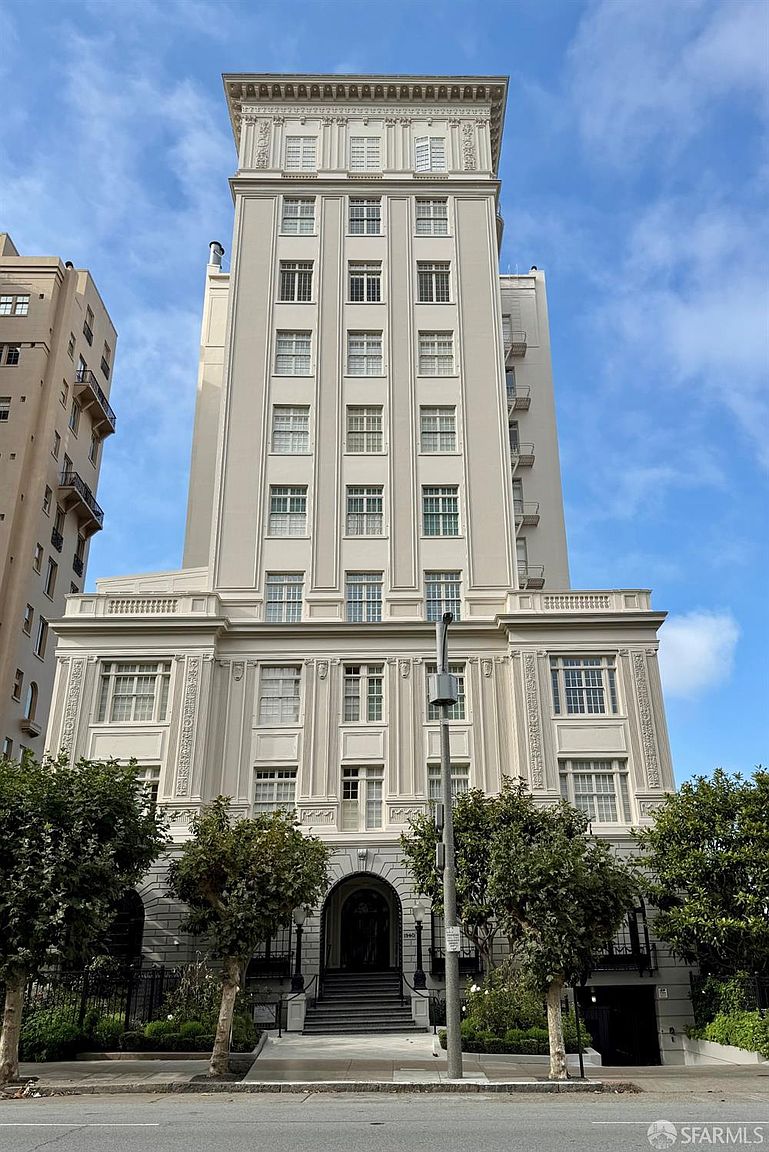 This image presents a direct, eye-level front view of a stately, multi-story historic apartment building with a symmetrical neoclassical facade. The structure features a light-colored stone exterior, a prominent arched entryway with steps leading up, and a series of evenly spaced windows that rise toward a decorative cornice at the roofline. Lush green trees frame the lower level, adding a touch of natural softness to the formal, urban architectural design.