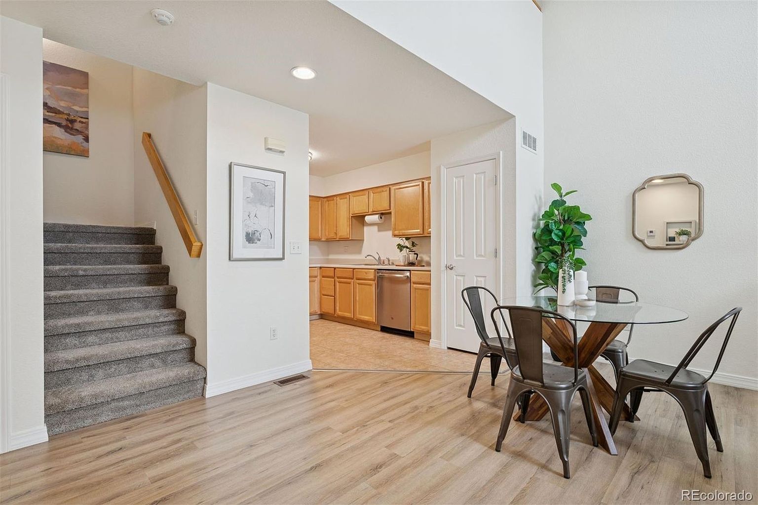 This interior shot showcases a dining area adjacent to a kitchen and stairs. The dining space features a round glass table with metal chairs, complemented by a decorative mirror and a potted plant. The flooring is light-colored wood, and the overall aesthetic is bright and inviting, creating a welcoming atmosphere for meals and gatherings.