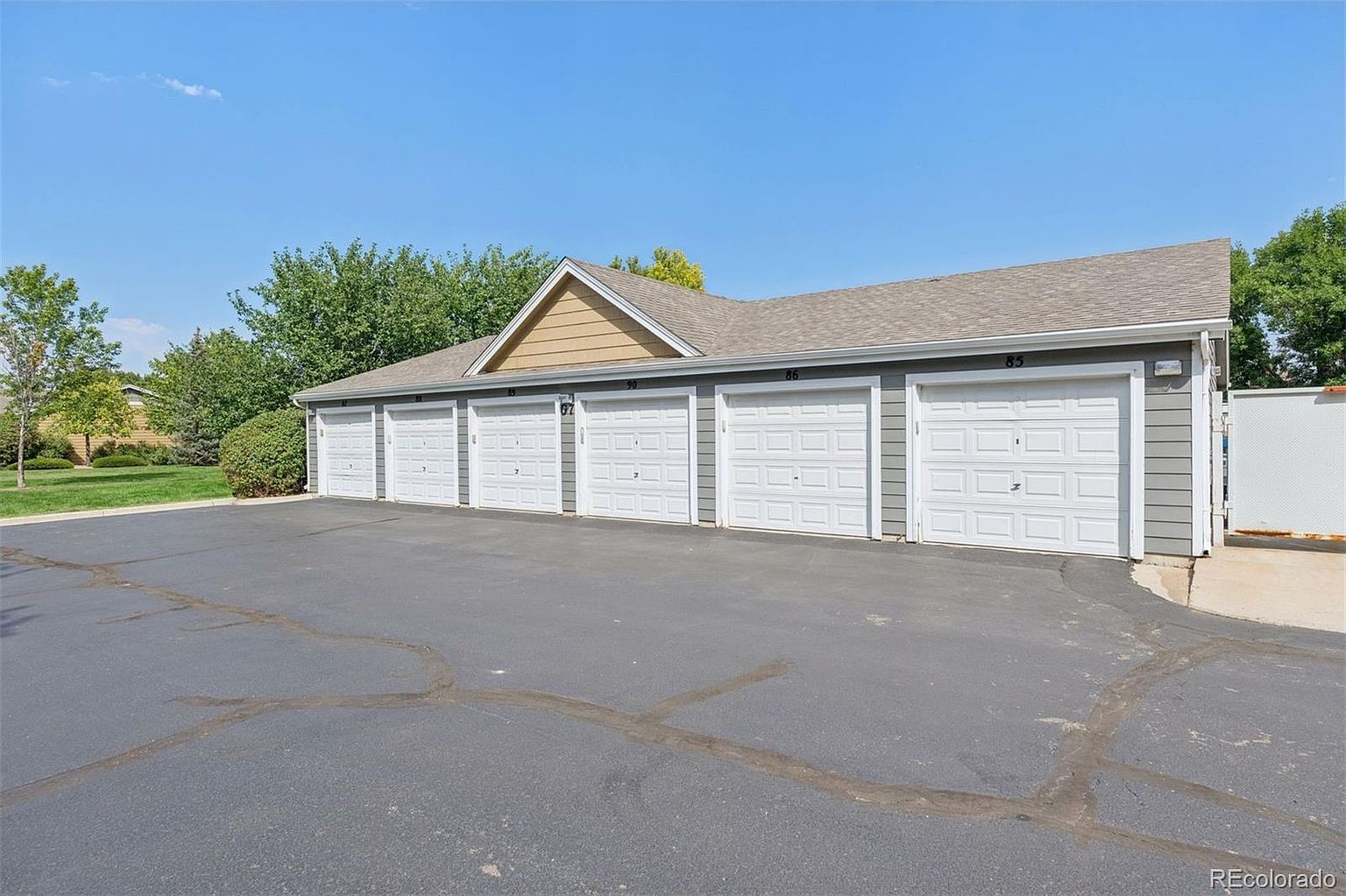 The image showcases a row of detached garages, likely serving a residential community. Each garage features a white door with a paneled design, set against a gray exterior. The garages are arranged linearly, with an asphalt driveway in front, under a clear blue sky, creating a neat and functional aesthetic.