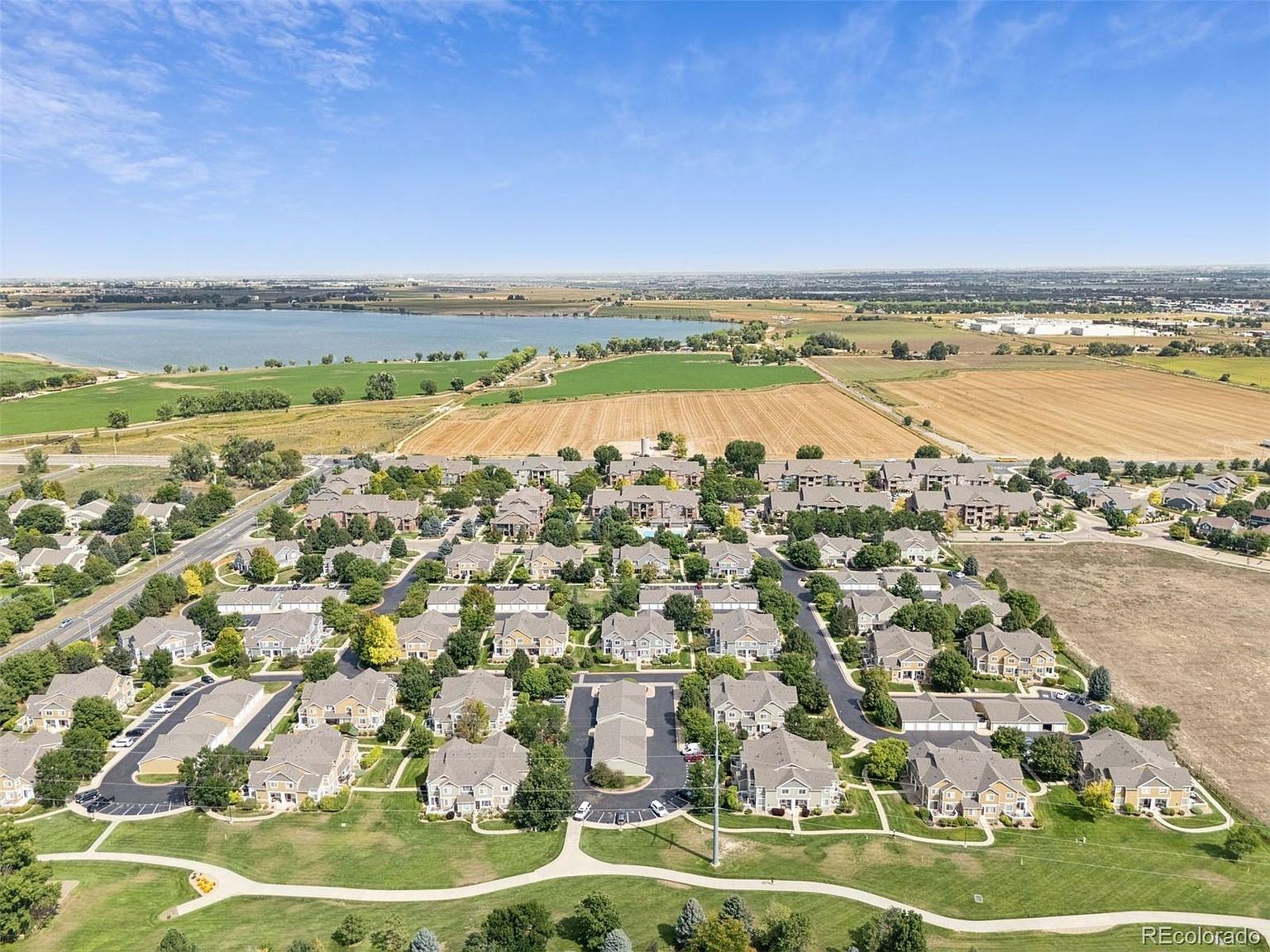 This aerial view showcases a well-planned residential community with neatly arranged houses, mature trees, and green spaces. A lake and agricultural fields are visible in the background, adding to the scenic appeal. The perspective highlights the community's layout and its integration with the surrounding natural environment.